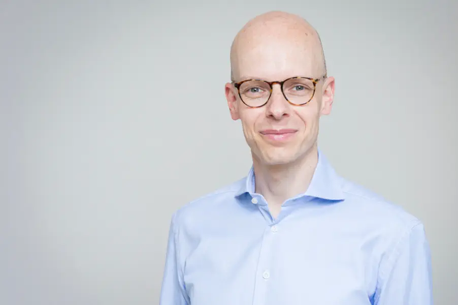 Sympathischer junger Mann ohne Haare. Er trägt ein hellblaues Hemd und eine große Hornbrille.