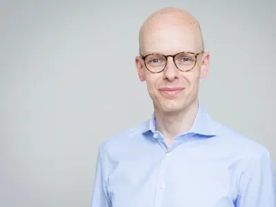 Sympathischer junger Mann ohne Haare. Er trägt ein hellblaues Hemd und eine große Hornbrille.