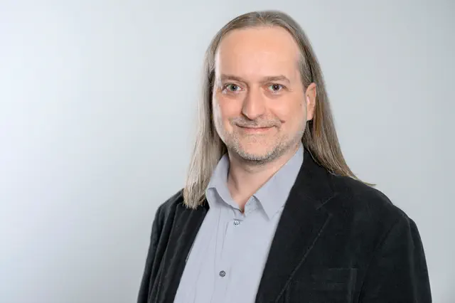 Freundlicher Mann mit langen  Haaren und Dreitagebart in hellem Hemd und dunklem Sakko.