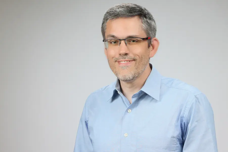 Freundlicher Mann mit kurzen grauen Haaren und Bartansatz mit Brille und hellem Hemd.