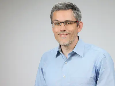 Freundlicher Mann mit kurzen grauen Haaren und Bartansatz mit Brille und hellem Hemd.